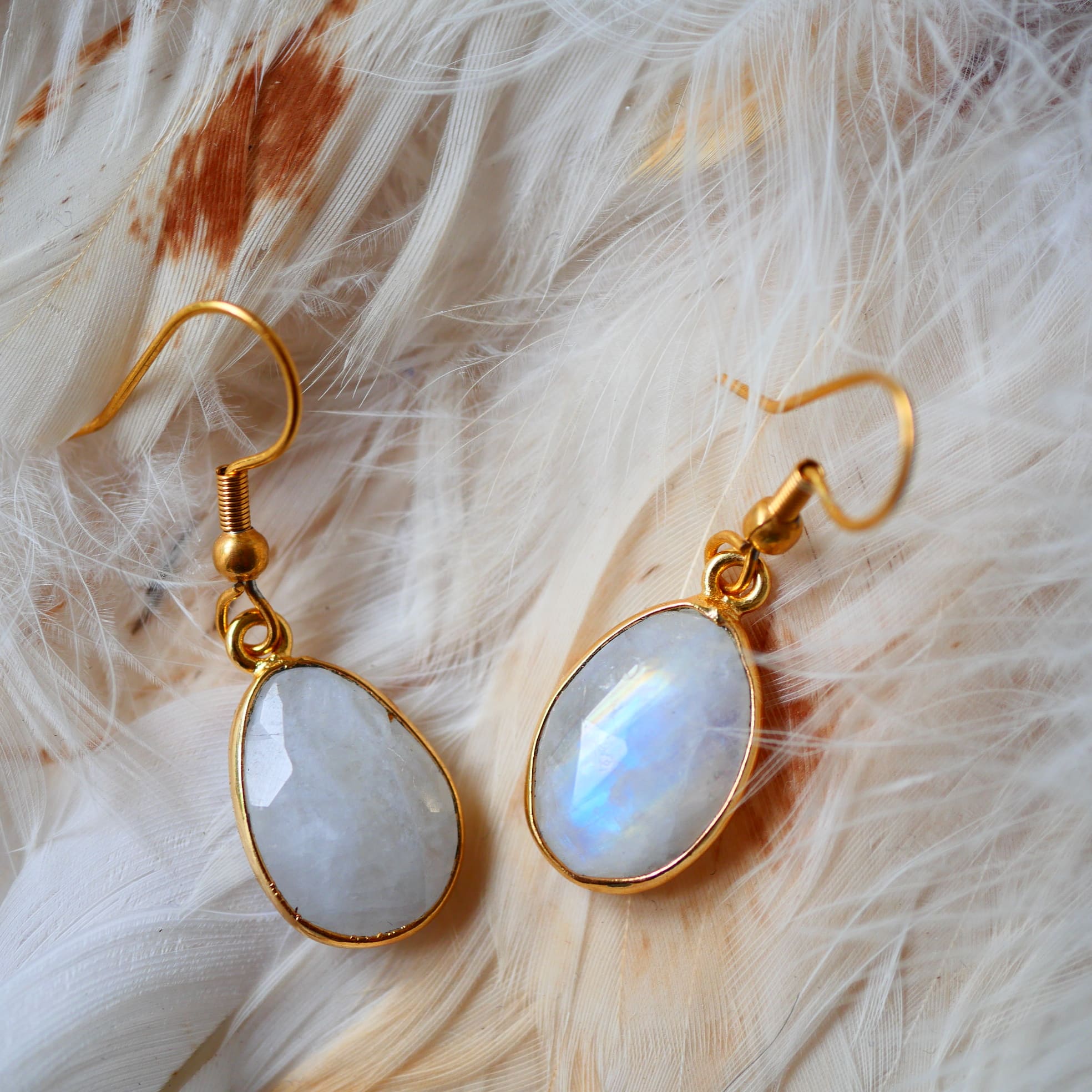 Boucles d'oreilles KAUMUDI Ginandger posées sur plumes – pierres de lune ovales facettées à reflets bleus irisés serties or, crochet doré, bijou pierres naturelles fait main à Paris
