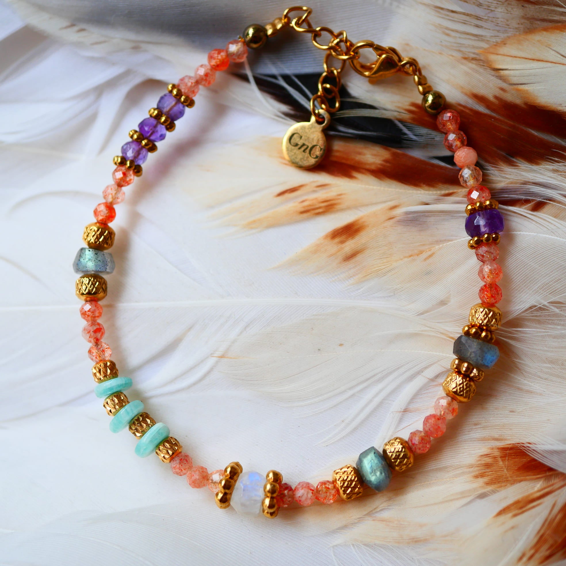 Bracelet SAMA Ginandger posé sur plumes blanches – pierres de soleil, améthystes, labradorites et amazonites, perles dorées ciselées, bijou pierres naturelles fait main à Paris
