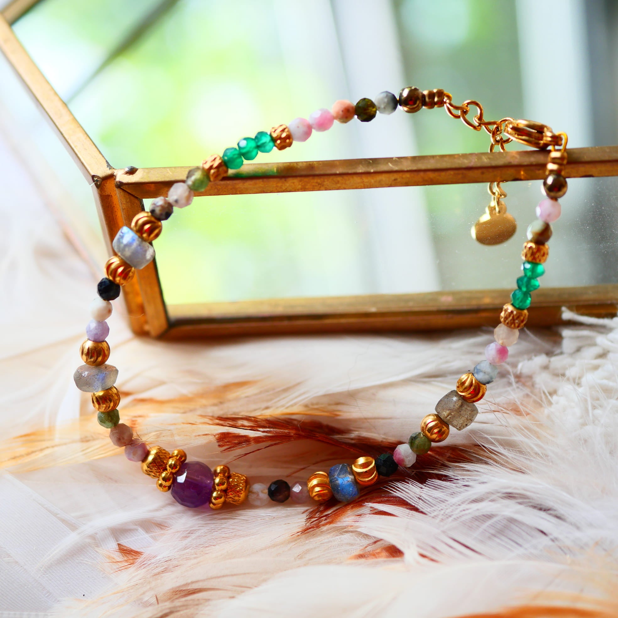 Bracelet THIRA Ginandger posé sur plumes et miroir doré – tourmalines multicolores, améthystes, labradorites et onyx verts, perles dorées, bijou pierres naturelles fait main à Paris
