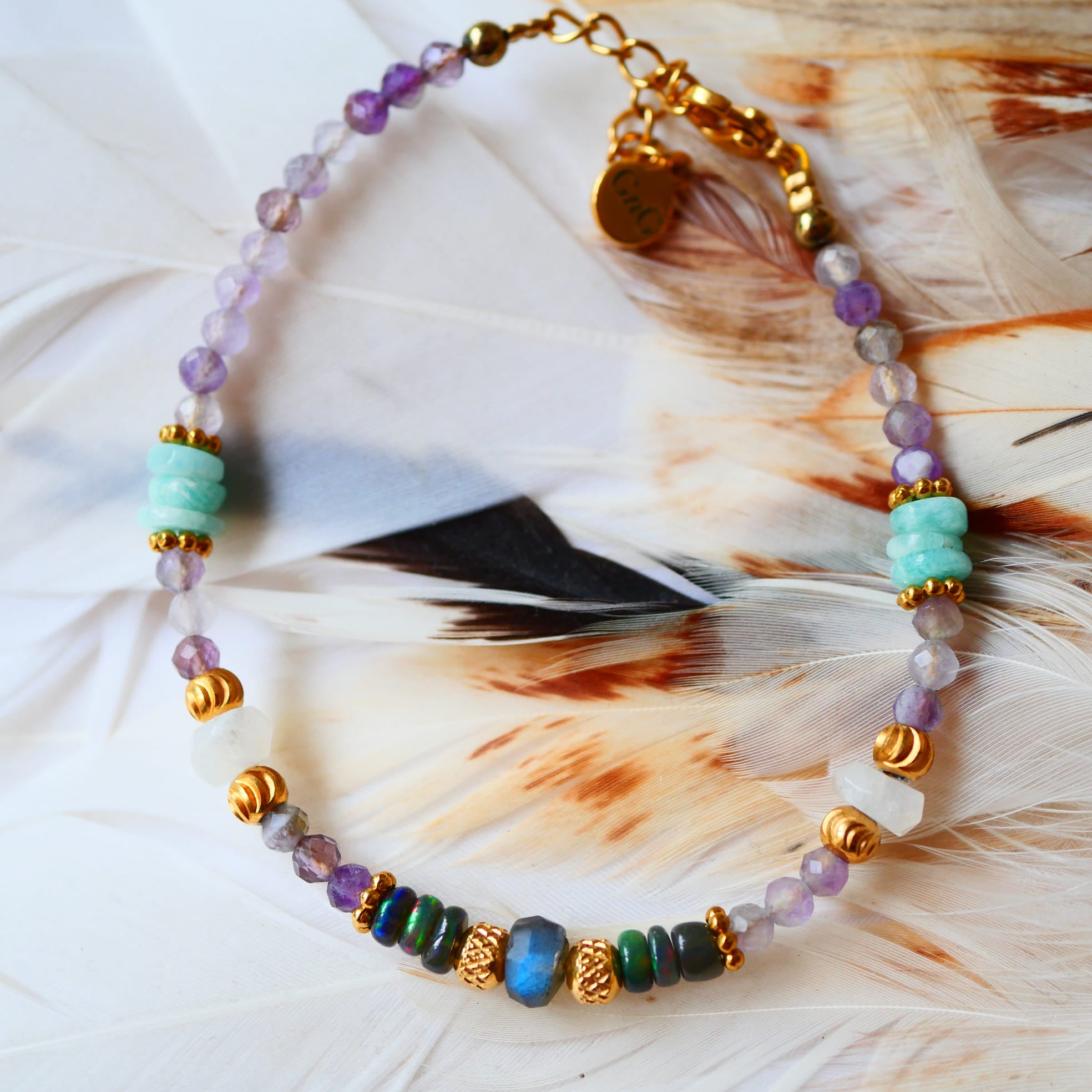 Bracelet SUKHA Ginandger posé sur plumes blanches – améthystes, amazonites, labradorites et onyx noirs irisés, perles dorées ciselées, bijou pierres naturelles fait main à Paris
