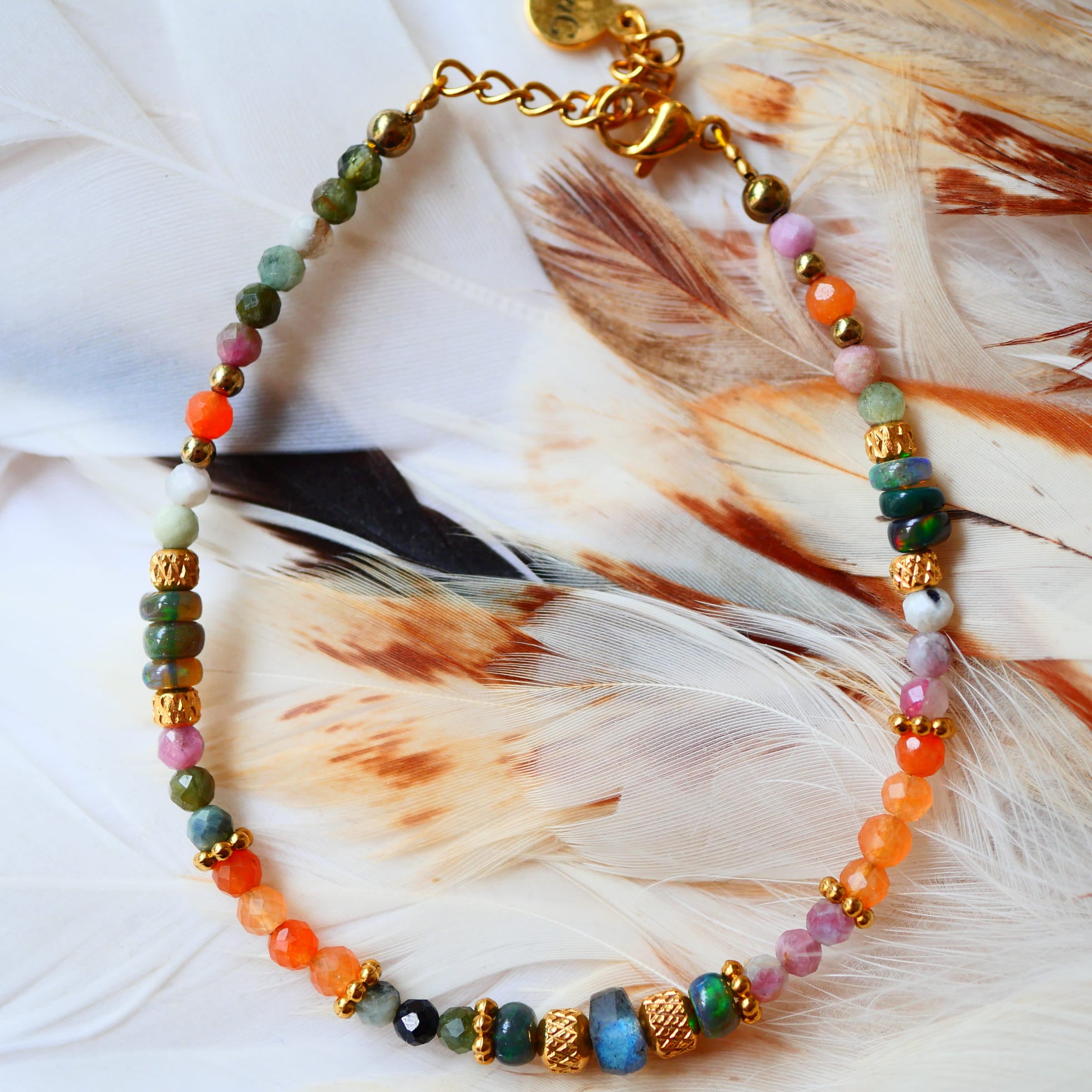 Bracelet VEERA Ginandger posé sur plumes blanches – tourmalines multicolores, carnélines et perles dorées, bijou pierres naturelles fait main à Paris
