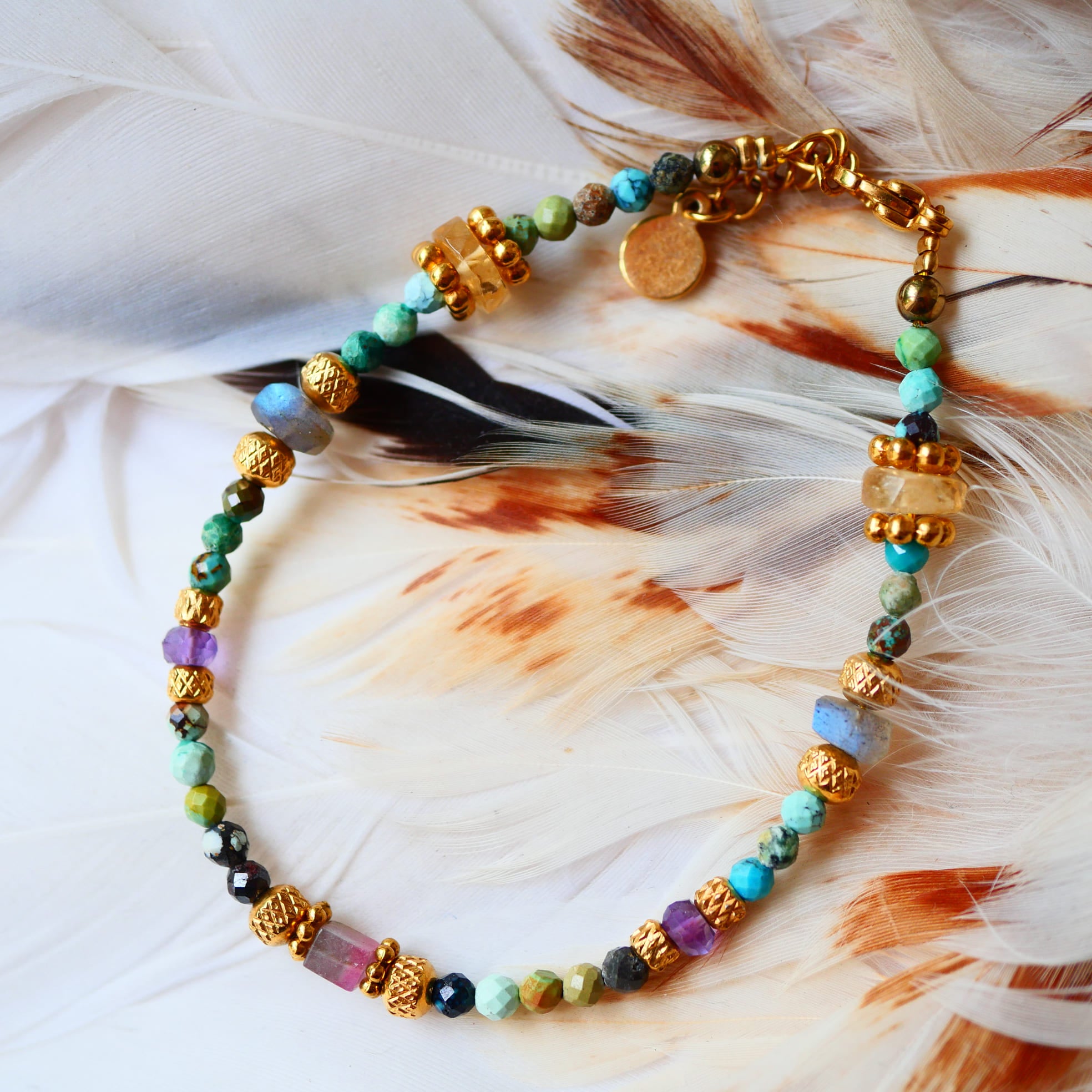 Bracelet YUKTI Ginandger posé sur plumes blanches – turquoises, améthystes, citrines et labradorites, perles dorées ciselées, bijou pierres naturelles fait main à Paris
