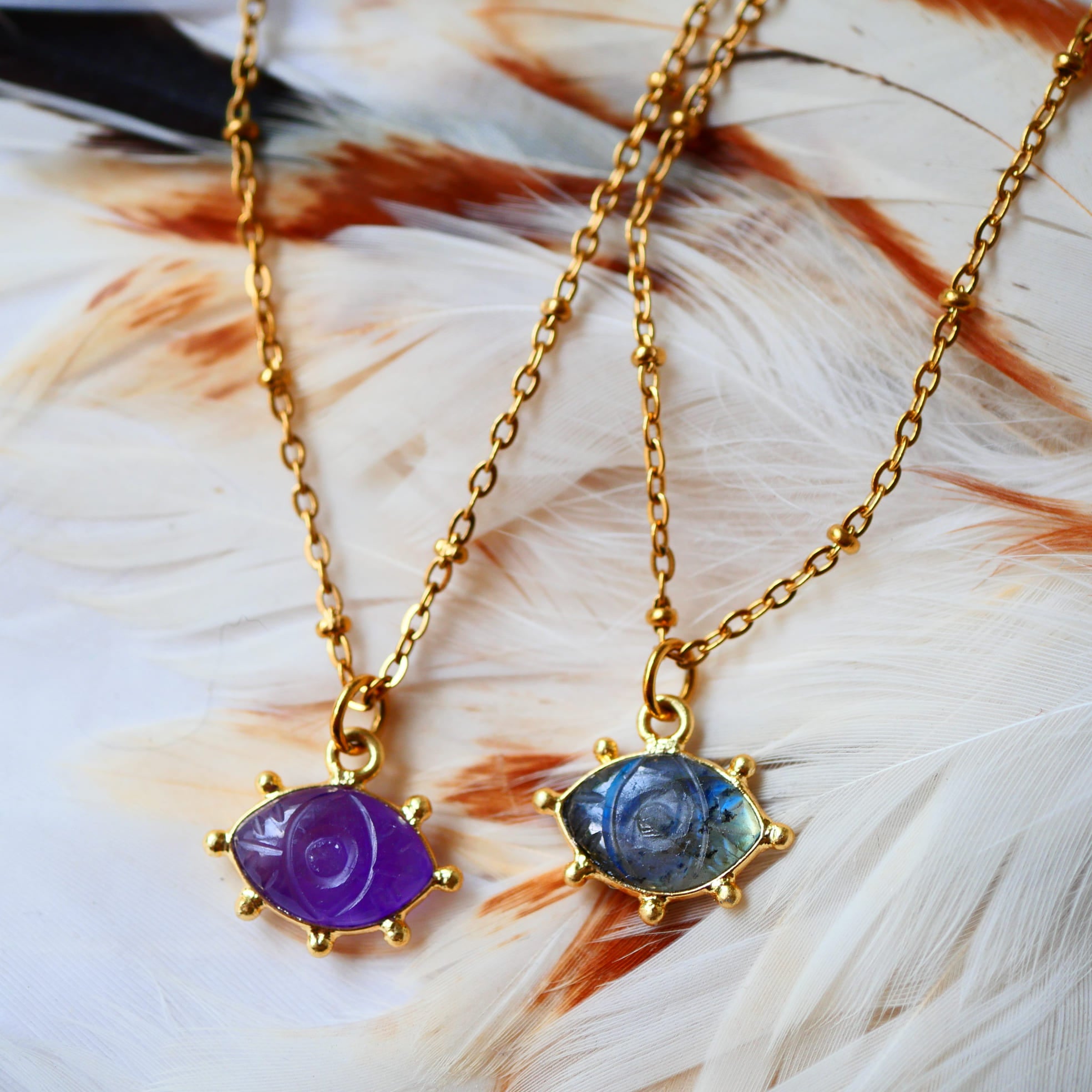 Collier AJNA Ginandger - pendentif œil améthyste violet et labradorite bleue sur chaîne dorée, fond plumes
