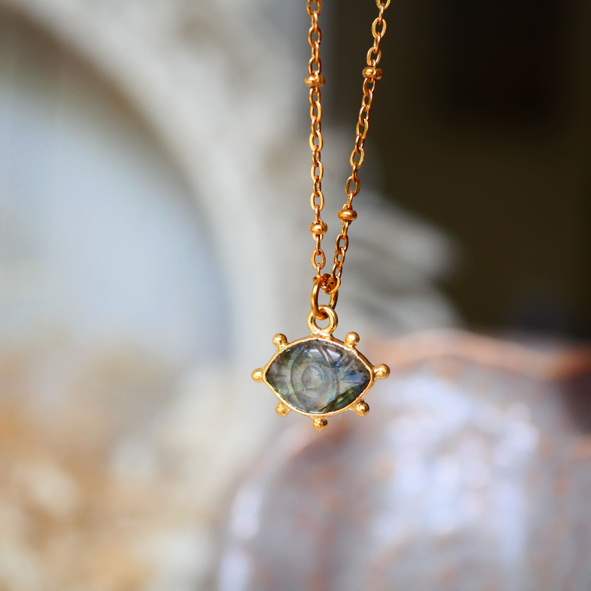 Collier AJNA Ginandger labradorite - pendentif œil sculpté bleu-gris serti or, suspendu sur fond de cristal
