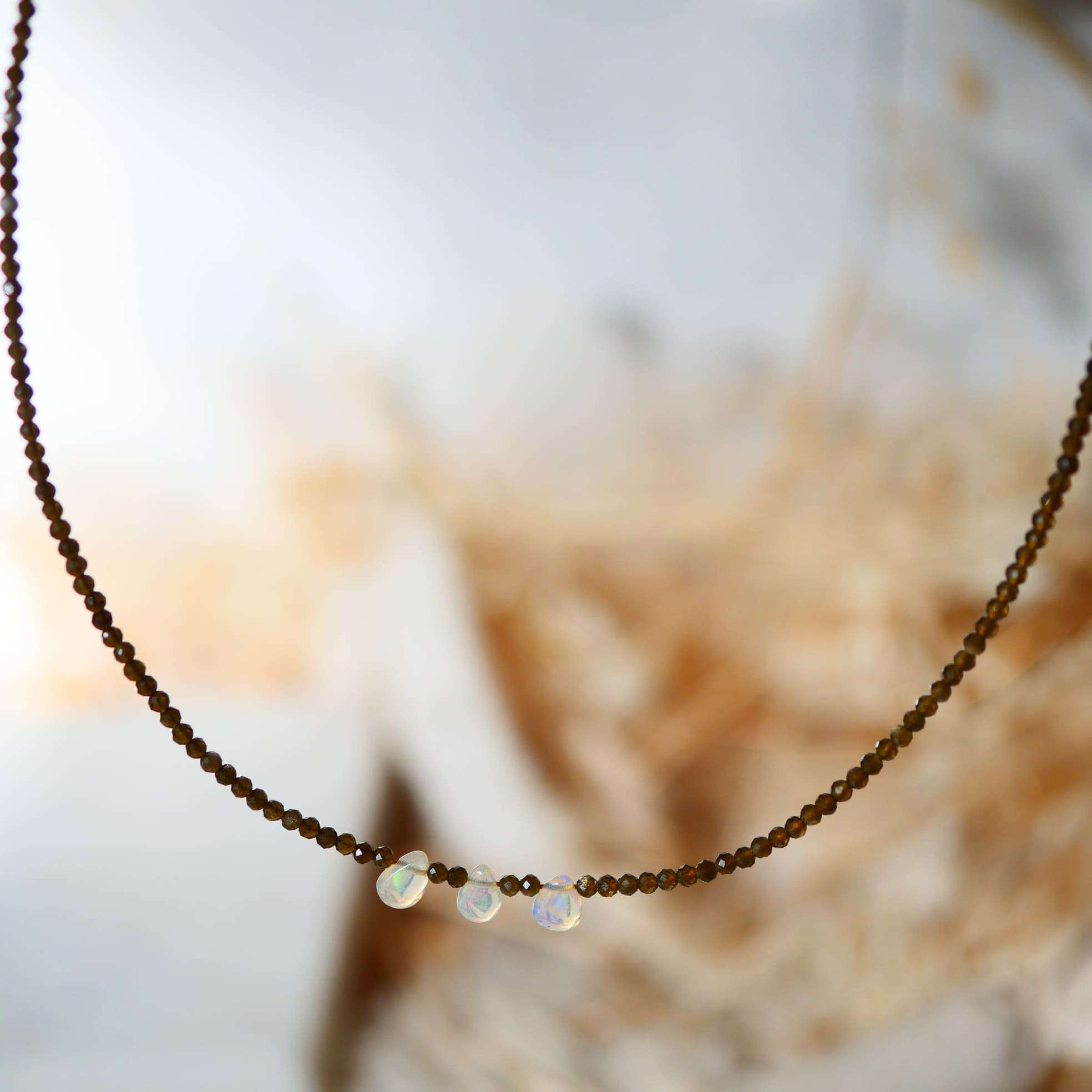 Collier ANTARA Ginandger suspendu - rang d'obsidienne dorée et gouttes d'opale irisées, fond floral naturel
