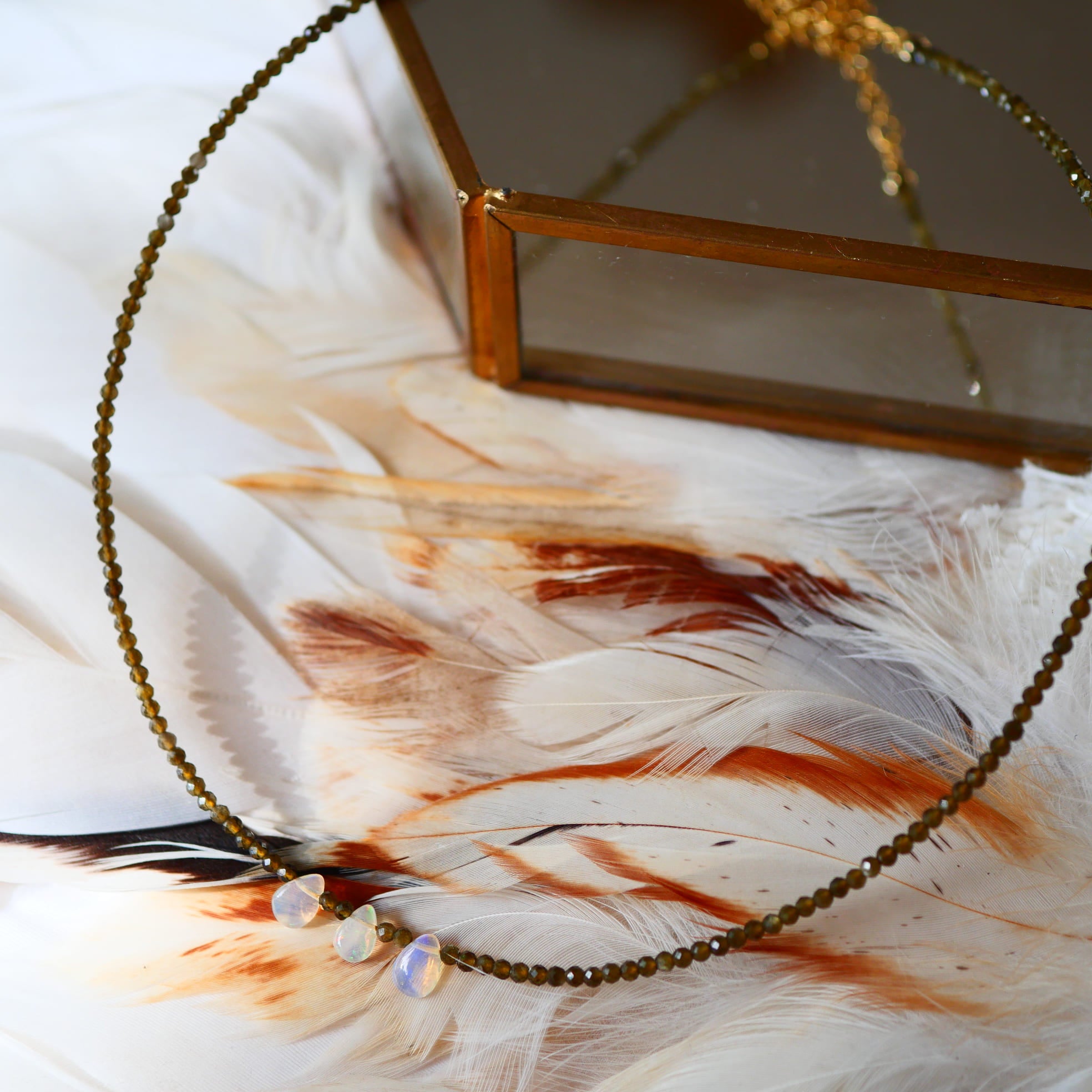 Collier ANTARA Ginandger - rang d'obsidienne et gouttes d'opale irisées posé sur plumes, écrin doré en fond
