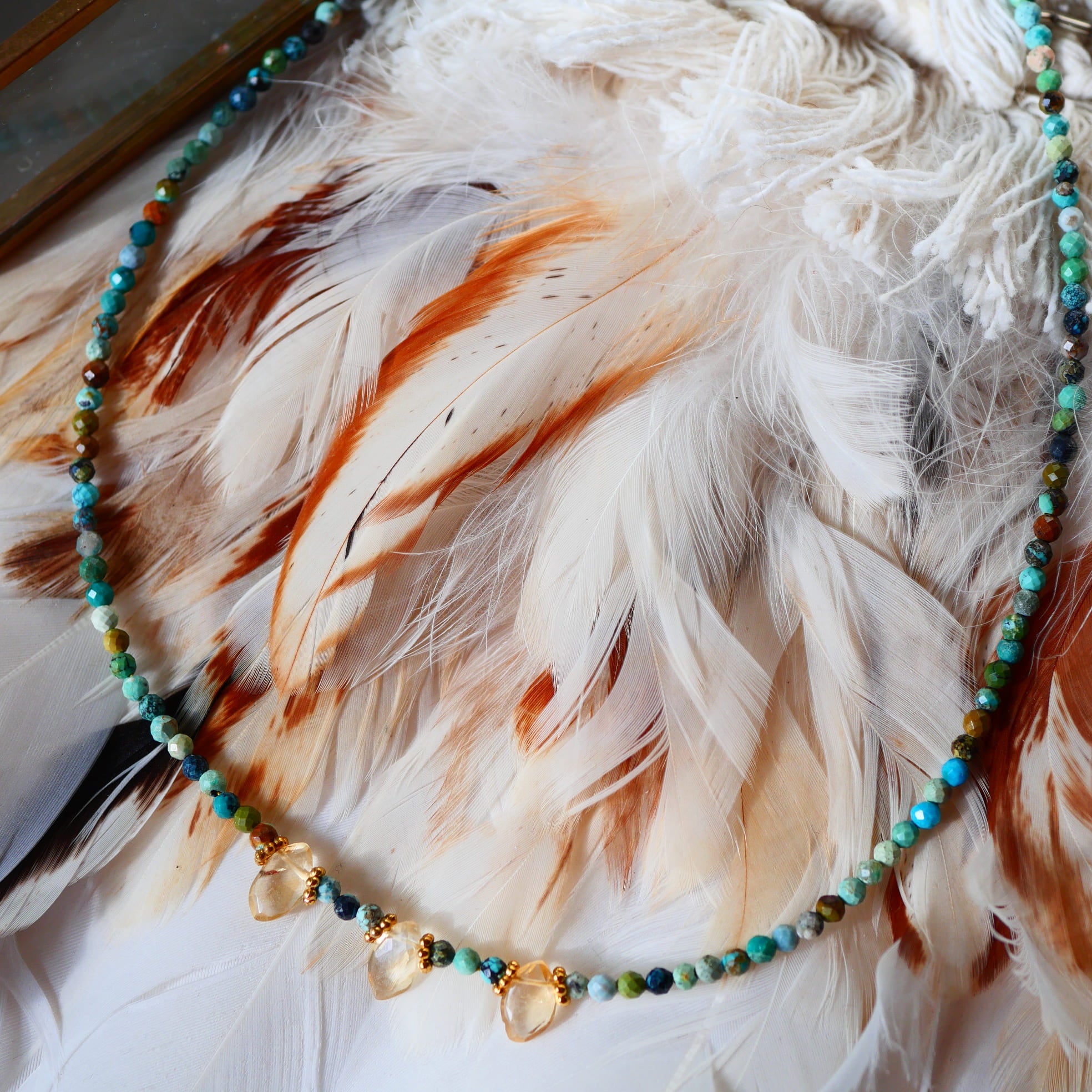 Collier DEEPTI Ginandger - rang de turquoises multicolores et pendentifs citrines sur chaîne dorée, fond plumes
