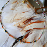Collier OJAS Ginandger posé sur plumes blanches – tourmalines multicolores, perles dorées et breloque plume, bijou pierres naturelles fait main à Paris
