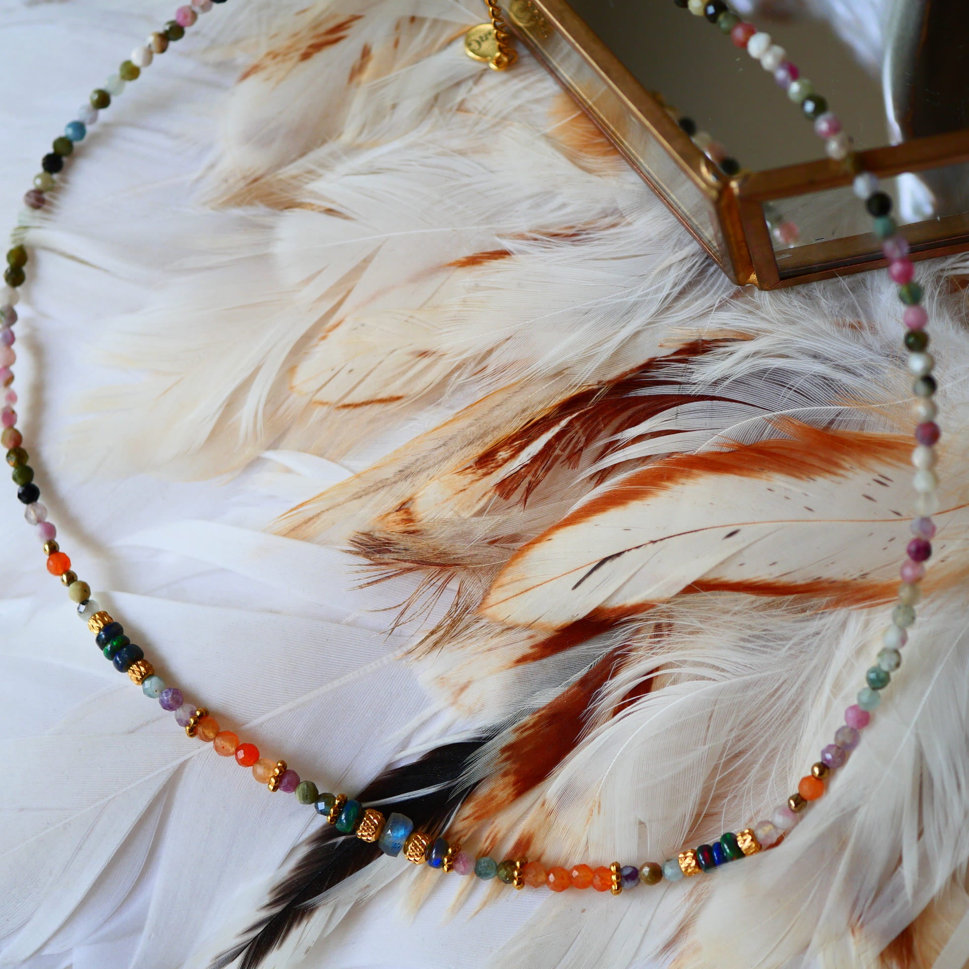 Collier OJAS Ginandger posé sur plumes blanches – tourmalines multicolores, perles dorées et breloque plume, bijou pierres naturelles fait main à Paris

